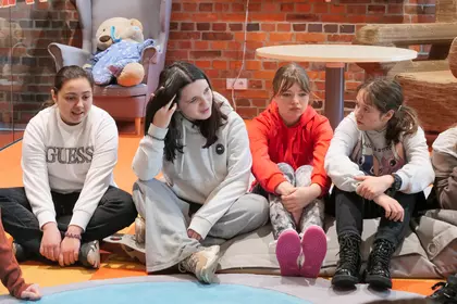 Uczniowie siedzą na podłodze i słuchają prowadzącej podczas zajęć.