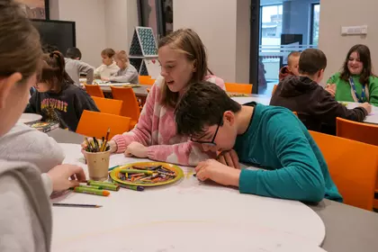 Dziecko rysuje przy stole, w tle inni uczestnicy zajęć.