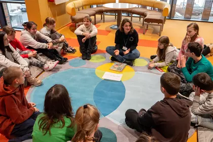 Uczestnicy warsztatów siedzą w kręgu na kolorowym dywanie.