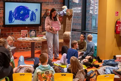 Zajęcia czytelnicze dla dzieci w bibliotece, prowadząca czyta książkę grupie słuchaczy.