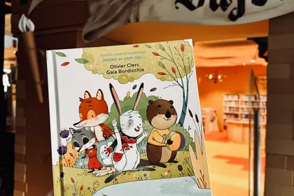 Książka dla dzieci z ilustracjami zwierząt trzymana na tle bibliotecznych regałów.