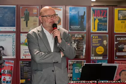 Mężczyzna w okularach mówi do mikrofonu na tle ściany z plakatami wydarzeń.