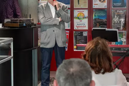 Prelegent w jasnej marynarce przemawia do publiczności, widoczne plecy słuchaczy.