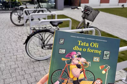 Książka „Tata Oli dba o formę” trzymana przed biblioteką i rowerami