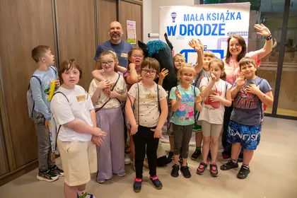 Grupa dzieci i prowadzący pozują przy planszy „Mała książka w drodze”.