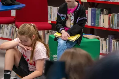 Dziewczynka z książką siedzi na pufie wśród uczestników wydarzenia.