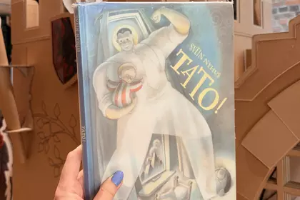 Dłoń trzyma książkę „Tato!” na tle kartonowego zamku w Oddziale dla Dzieci.