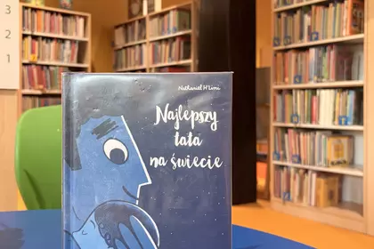 Książka „Najlepszy tata na świecie” ustawiona na niebieskim stoliku wśród bibliotecznych regałów.