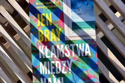 Książka „Kłamstwa między nami” leżąca na drewnianym leżaku w promieniach słońca.