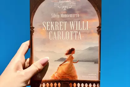 Dłoń trzyma książkę „Sekret willi Carlotta” na tle błękitnego nieba.