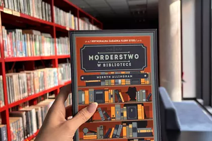 Książka „Morderstwo w bibliotece” trzymana na tle regałów bibliotecznych.