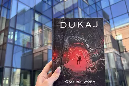 Dłoń trzyma książkę „Oko potwora” na tle nowoczesnej szklanej elewacji.