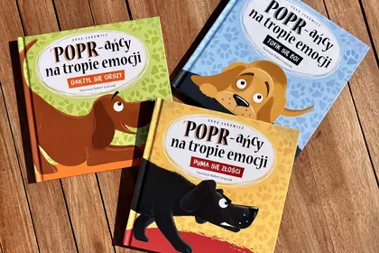 Trzy książki z serii „POPR-ańcy na tropie emocji” autorstwa Anny Sakowicz ułożone na drewnianym blac