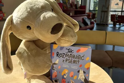 Pluszowy pies obejmuje książkę „Jak rozmawiać z psem?” ustawioną na stoliku w Oddziale dla Dzieci i 