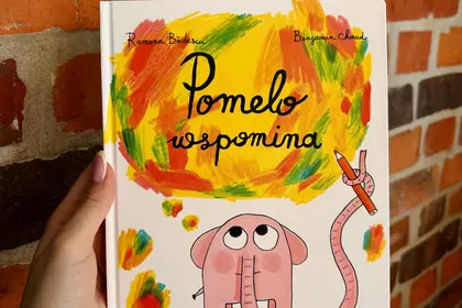 Książka „Pomelo wspomina” trzymana w dłoni na tle ceglanej ściany.