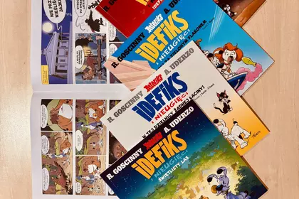 Cztery tomy komiksów o Idefiksie rozłożone na blacie obok otwartego egzemplarza pokazującego ilustra