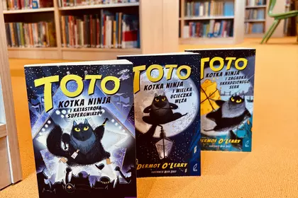 Trzy tomy serii „Toto. Kotka ninja” ustawione na pomarańczowej wykładzinie pomiędzy regałami bibliot