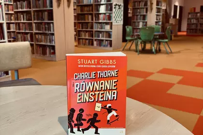 Powieść „Charlie Thorne i równanie Einsteina” Stuarta Gibbsa ustawiona na stoliku w bibliotece, w tl