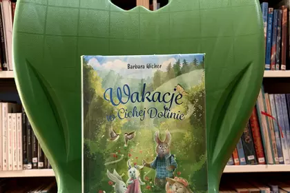 Książka „Wakacje w Cichej Dolinie” ustawiona na zielonym krześle w bibliotece.