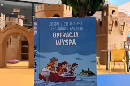 Książka „Operacja Wyspa” na tle dziecięcej strefy zabaw w bibliotece.