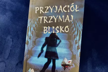 Książka „Przyjaciel trzymaj blisko” na granatowym fotelu.