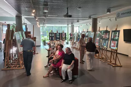Goście siedzą na kanapie i oglądają wystawę kolorowych grafik na sztalugach w galerii.