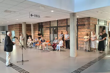Starszy mężczyzna przemawia do mikrofonu w nowoczesnym holu przed grupą słuchaczy.