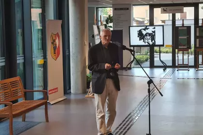 Mężczyzna w beżowej marynarce przemawia do mikrofonu w przeszklonym holu biblioteki.