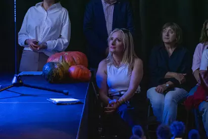 Uczestnicy Narodowego Czytania siedzą obok sceny udekorowanej dyniami.